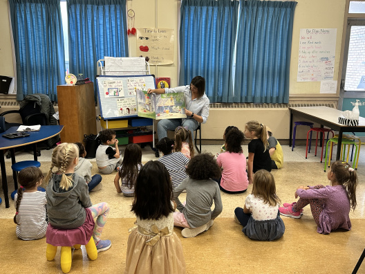 Children sitting on the floor, listening to a teacher read a book